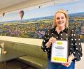 Foto van een vrouw die glimlachend een ingelijst certificaat vast houdt over "Dementievriendelijke ontmoetingsplek". Ze staat in een kantoorruimte met een grote wandfoto van een landschap met luchtballonnen.